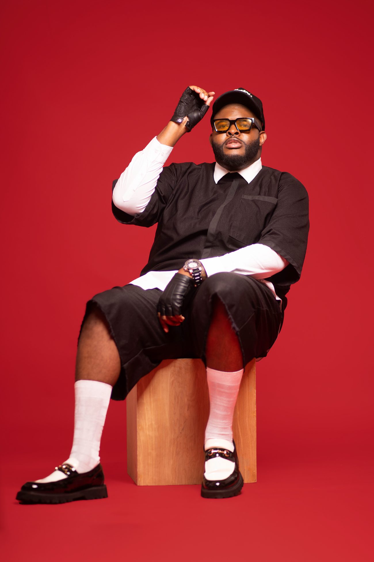 Jay Washington seated on a wooden block against a red studio wall, in black and white with yellow-tinted glasses